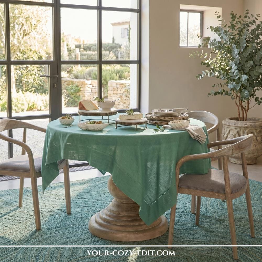 A round wooden pedestal table is elegantly draped with a green linen tablecloth and styled with artisanal serving platters and small white bowls. The setup sits on a large, textured circular rug in a deep seafoam green, surrounded by light wood dining chairs with grey cushions. In the background, large glass-paned doors look out onto a sunlit Mediterranean-style courtyard with stone walls. A tall eucalyptus tree in a decorative stone planter completes the serene, organic modern aesthetic of the room.