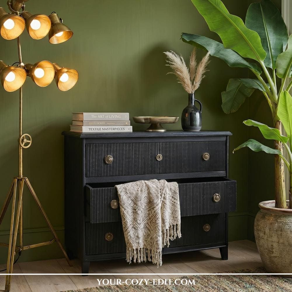 A sophisticated interior scene featuring a black fluted dresser with brass knobs against a deep olive green wall. To the left, a multi-head brass floor lamp provides warm illumination, while a large banana leaf plant adds a lush, natural element. The dresser is styled with curated books and a ceramic vase, creating a serene and refined atmosphere. This composition perfectly balances moody tones with warm metallic accents.