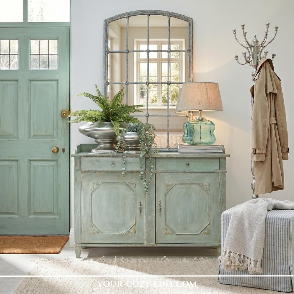 A sun-drenched entryway features a vintage-style sideboard in a muted mint green, paired with a matching paneled door and a neutral woven rug. A large, arched window-style mirror hangs above the cabinet, reflecting light alongside a silver coat rack and a striped ottoman. On the sideboard, a lush fern in a metallic planter and a seafoam glass lamp add organic texture and a warm glow. The overall design creates a harmonious blend of classic character and contemporary freshness through a soft green color scheme.