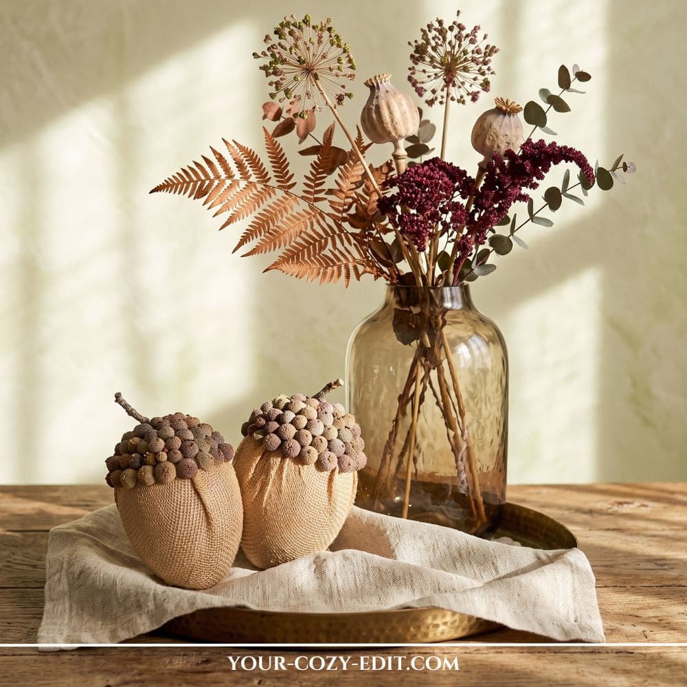 Two DIY oversized burlap acorns with textured caps made of natural cones, displayed on a gold tray next to an amber glass vase filled with dried ferns and poppy pods on a wooden table.
