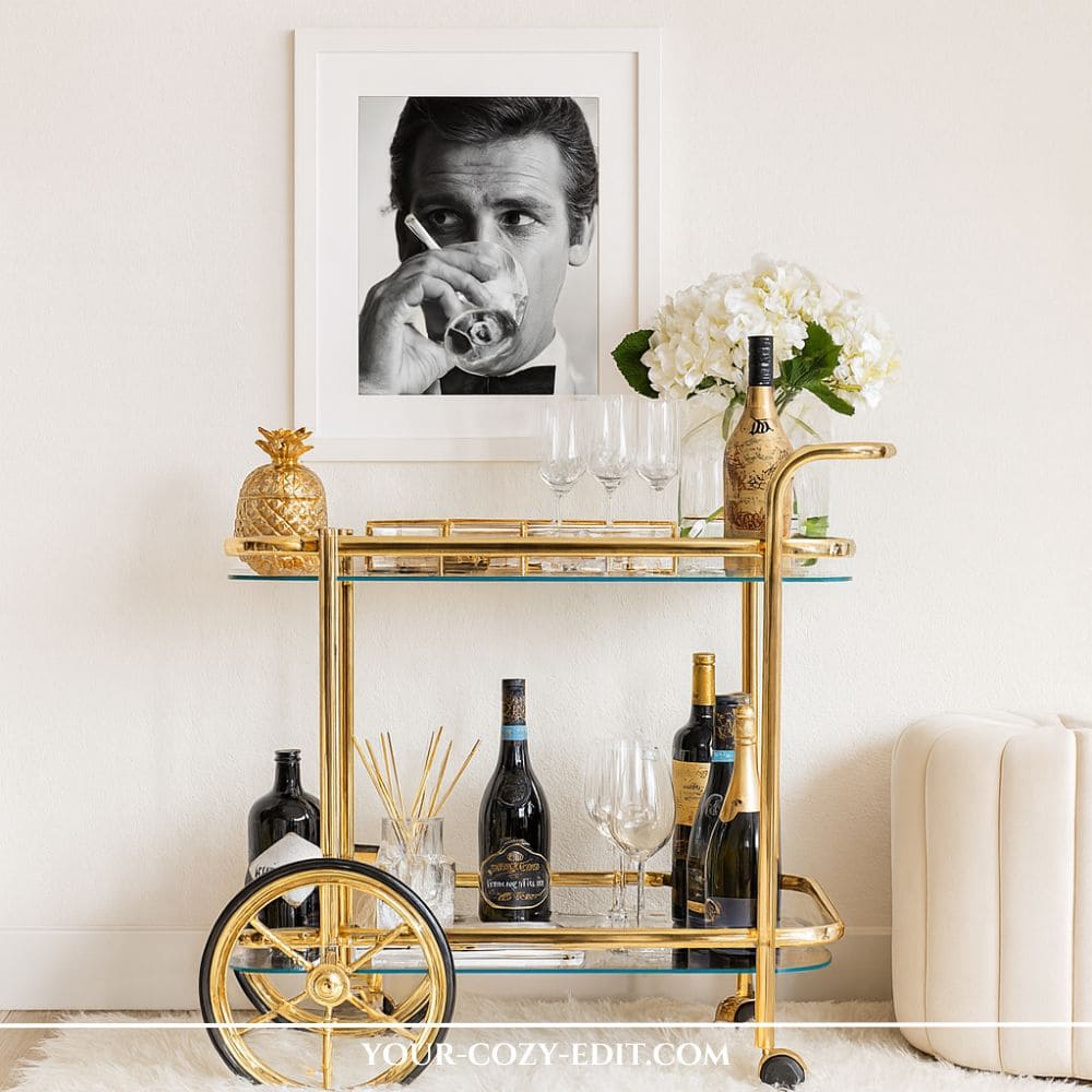shiny gold brass bar cart with large wheels styled with a framed black and white portrait of a man drinking a cocktail, a gold pineapple container, champagne flutes, white hydrangeas, and wine bottles against a white wall