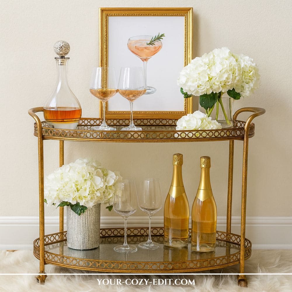 antique gold oval bar cart styled with white hydrangeas in vases, a framed peach cocktail illustration, a crystal decanter, wine glasses, and champagne bottles, anchored on a fluffy white faux fur rug